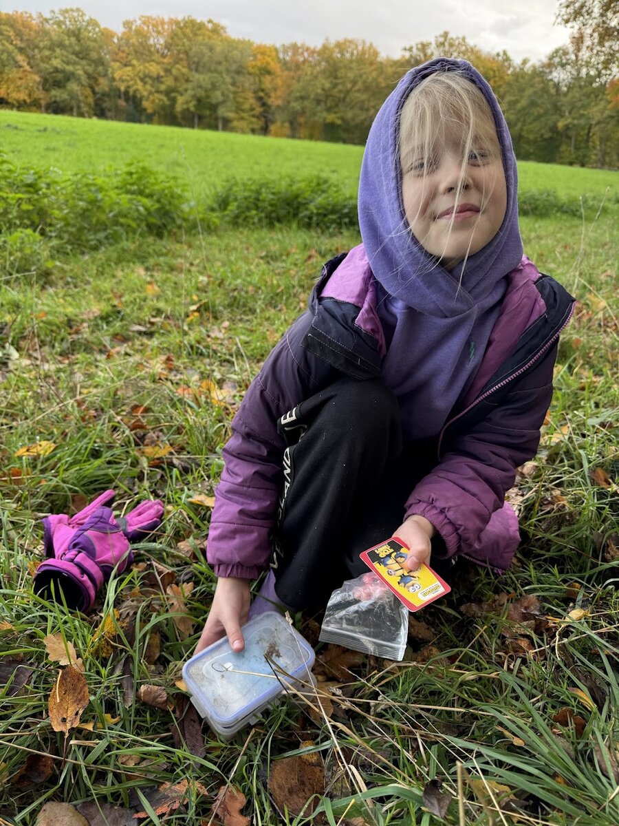 A child in a purple hooded sweatshirt crouching in a grassy field, holding a small clear plastic container and a Minions card, with pink gloves on the ground nearby
